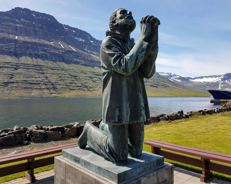 The monument for fishermen who drowned in Eskifjöður.  In the background, there is the mountain Hólmatindur. The sun is shining, and it is a beautiful day. 