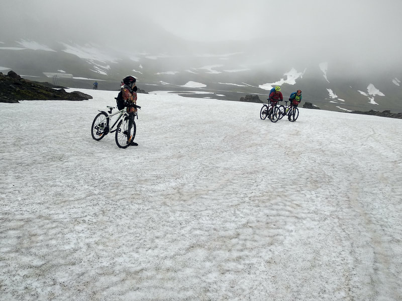 Three people with mountain bikes crossing a field of snow in Oddskarð. 