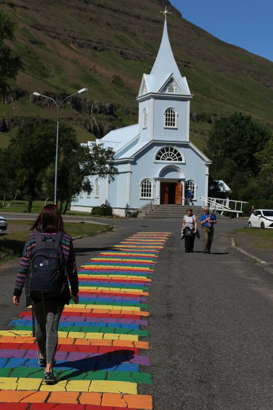 A young woman walks towards the blue church in Seyðisfjörður. The street she is walking is the rainbow street, painted in the gay pride colors. 