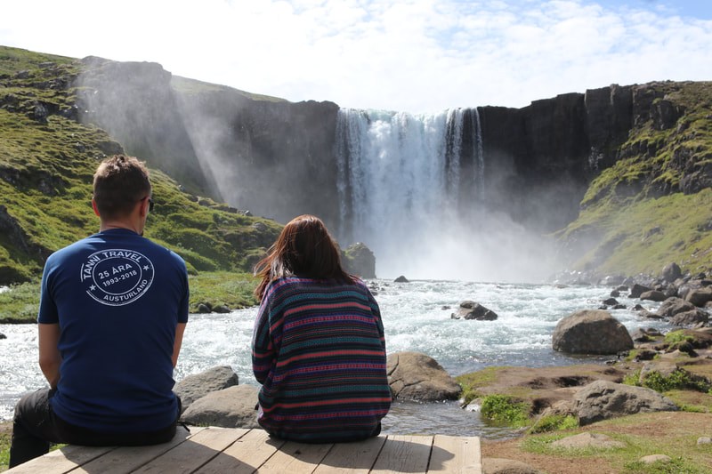 A man and a woman sitting in front of the waterfall Gufufoss in Seyðisfjörður. 